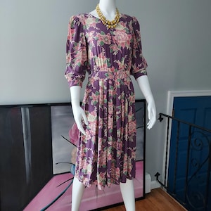 Puede incluir: Un vestido floral morado con cintura fruncida y mangas abullonadas. El vestido tiene un estampado floral rosa y blanco.