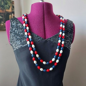 May include: A long, layered necklace featuring red, white, and blue oval beads. The necklace is displayed on a black dress with sequin detailing on the straps. The beads are strung together, creating a patriotic colour scheme.