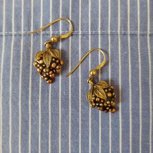 Puede incluir: Un par de pendientes colgantes dorados. Cada pendiente presenta un racimo de uvas con hojas detalladas. Los pendientes están colocados sobre un fondo a rayas azules y blancas.