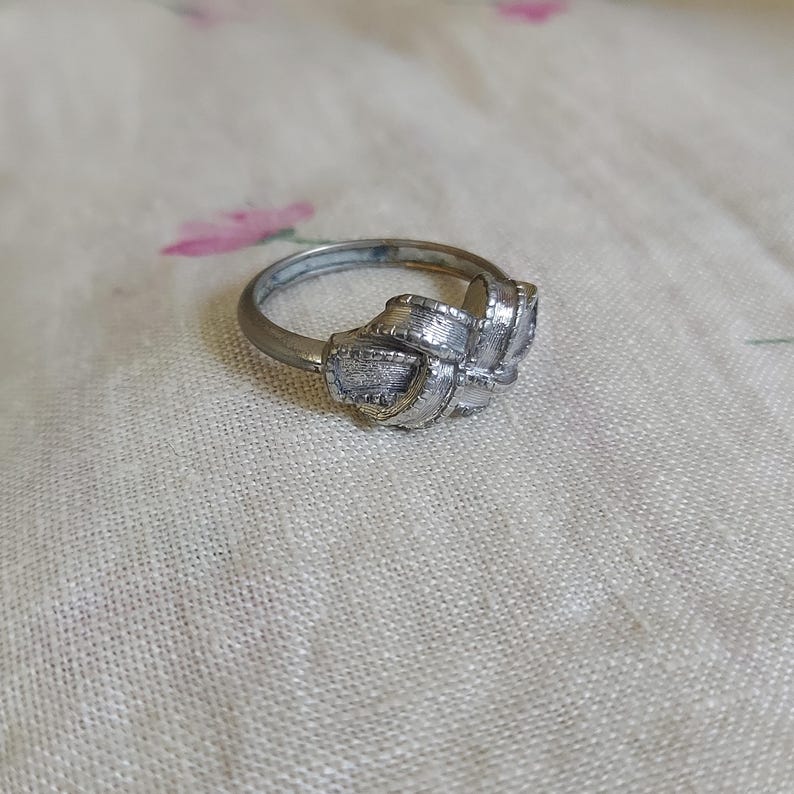 May include: A silver-toned metal ring featuring a central knot design. The band is smooth and circular, while the focal point consists of textured, overlapping loops that resemble a tied ribbon or a decorative knot with a metallic finish.