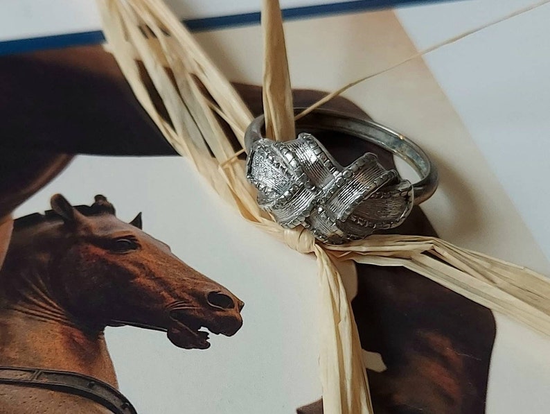 May include: A silver-toned metal ring featuring a textured, knotted ribbon design. The surface of the ring displays a brushed metallic finish with intricate cross-hatched detailing across the central knot element, creating a dimensional appearance.