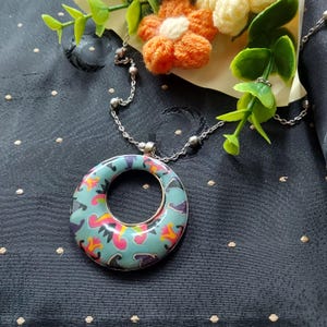 May include: A circular pendant necklace featuring a light blue base with a colorful pattern of pink, yellow, and dark purple abstract shapes. The pendant hangs from a thin silver-toned metal chain with small spherical beads spaced along the length.