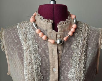 May include: A beaded necklace featuring round and teardrop-shaped peach-colored beads interspersed with metallic silver spherical and elongated beads. The necklace is displayed on a mannequin wearing a beige sleeveless top with lace trim.