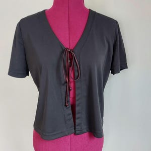 May include: A black short-sleeve top with a V-neckline and a tie closure. The top is made of a smooth fabric and has a simple, classic design. The tie is a dark brown color.