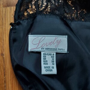 May include: Close-up of a black dress with a lace neckline. The label reads "Lovely by Adrianna Papell" with size information: US/CAN 8, EU 40, UK 12, GER 38, AUS 12. The dress is made in China.