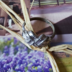 May include: A silver-toned metal ring featuring a textured, woven-style knot design at the center. The band is thin and circular, with a polished finish that reflects light, showcasing a detailed, crisscross pattern on the front decorative element.