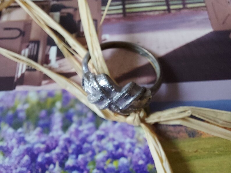 May include: A silver-toned metal ring featuring a textured, woven-style knot design at the center. The band is thin and circular, with a polished finish that reflects light, showcasing a detailed, crisscross pattern on the front decorative element.