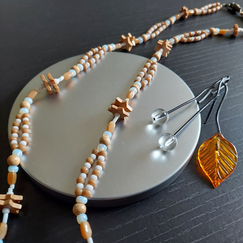 K&ouml;nnte beinhalten: Eine braune und wei&szlig;e Perlenkette mit h&ouml;lzernen sternf&ouml;rmigen Anh&auml;ngern. Die Kette wird auf einem silbernen Tablett mit zwei Paar Ohrringen und einem kleinen orangefarbenen Blattanh&auml;nger pr&auml;sentiert.