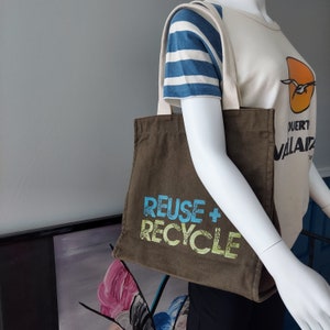 Puede incluir: Una bolsa de lona marrón con el texto "REUSE + RECYCLE" en letras azules y verdes.