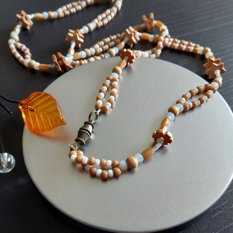 K&ouml;nnte beinhalten: Eine zweireihige Halskette aus braunen und wei&szlig;en Perlen mit einem kleinen Metallanh&auml;nger. Die Perlen sind blumenf&ouml;rmig und auf einer braunen Schnur aufgef&auml;delt.