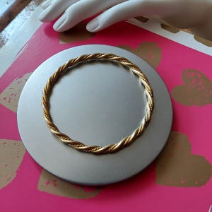 May include: A gold-toned metal bangle bracelet featuring a continuous twisted rope design. The circular bracelet has a polished finish and a uniform thickness throughout its entire circumference, creating a simple and classic jewellery piece.