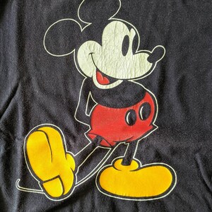 以下が含まれることがあります： 黒のTシャツに、赤と黒の衣装を着たミッキーマウスの漫画の画像がプリントされています。ミッキーマウスは笑顔で、黄色の靴を履いています。