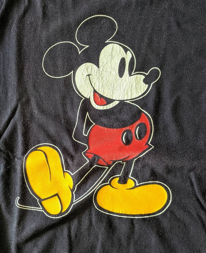 以下が含まれることがあります： 黒のTシャツに、赤と黒の衣装を着たミッキーマウスの漫画の画像がプリントされています。ミッキーマウスは笑顔で、黄色の靴を履いています。