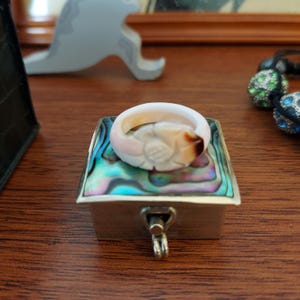 May include: A carved shell ring with a smooth, pale pink and white surface featuring a dark brown accent, resting on a small square box with an iridescent abalone shell inlay top and a silver-toned metal base with a central latch mechanism.