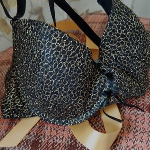 May include: Black bra with gold lace detailing. The bra has adjustable straps and a detailed lace pattern. It is displayed on a wooden hanger against a patterned background.