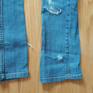 May include: Close-up of light blue denim jeans with distressed details. The jeans have ripped sections and frayed edges, with visible stitching in a contrasting color. The fabric shows signs of wear and tear, suggesting a vintage or worn aesthetic.
