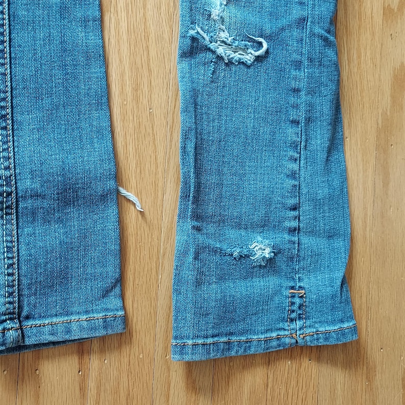May include: Close-up of light blue denim jeans with distressed details. The jeans have ripped sections and frayed edges, with visible stitching in a contrasting color. The fabric shows signs of wear and tear, suggesting a vintage or worn aesthetic.