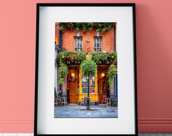 May include: A vintage-style pub with a green awning and the words "21  PARR - 21" above the entrance. The pub has two large windows with yellow frames and a wooden door. There are hanging plants and flowers in front of the pub.