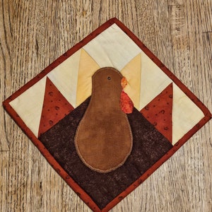 May include: A brown and white quilted turkey-shaped wall hanging with a red wattle. The turkey is made of fabric scraps and has a brown felt body.