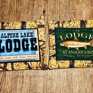 May include: Two square fabric pot holders with lodge-themed designs. One features "Alpine Lake Lodge" with "Hike, Swim, Fish, Canoe, Wildlife" text. The other says "The Lodge at Angler's Bay" with a fish illustration.