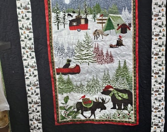 May include: A black and white quilted throw featuring a winter camping scene with a camper, a tent, a moose, a bear, and a snowy forest. The border is a white and green pattern with small houses.