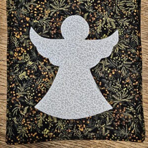 May include: A white angel cutout on a black fabric background with a green and gold floral pattern.