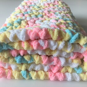 May include: A folded crocheted baby blanket in pastel colors of pink, yellow, and blue.