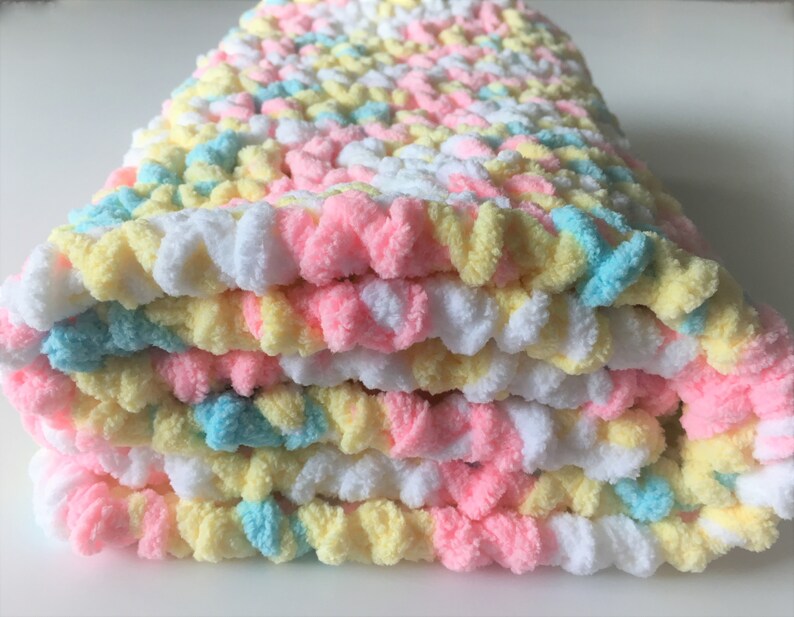 May include: A folded crocheted baby blanket in pastel colors of pink, yellow, and blue.
