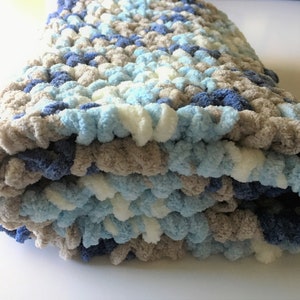 May include: A soft, crocheted baby blanket in shades of blue, white, and gray. The blanket is folded in thirds and has a fluffy texture.