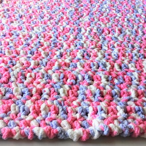 Puede incluir: Una suave manta de beb&eacute; tejida a crochet en rosa, blanco y lavanda. La manta est&aacute; hecha con un hilo grueso y tiene un aspecto texturizado y esponjoso.