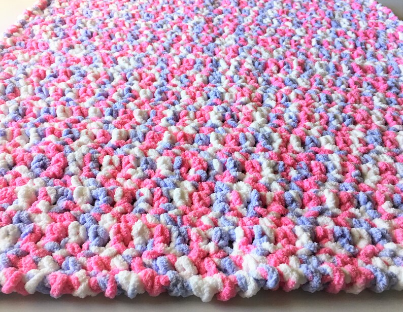 Puede incluir: Una suave manta de beb&eacute; tejida a crochet en rosa, blanco y lavanda. La manta est&aacute; hecha con un hilo grueso y tiene un aspecto texturizado y esponjoso.