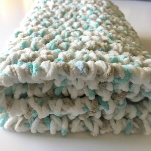 Puede incluir: Una suave manta de beb&eacute; tejida a crochet en blanco, beige y azul claro. La manta est&aacute; doblada en tres partes y tiene una textura esponjosa.