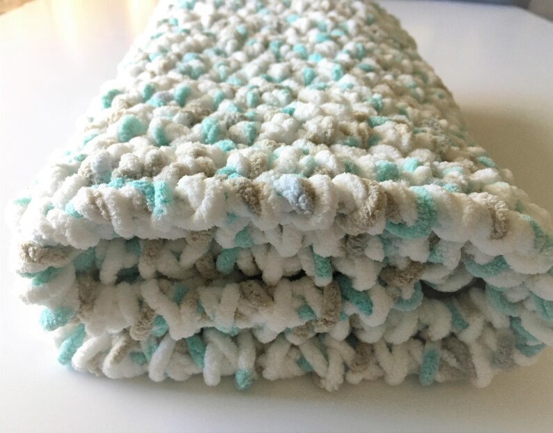 Puede incluir: Una suave manta de beb&eacute; tejida a crochet en blanco, beige y azul claro. La manta est&aacute; doblada en tres partes y tiene una textura esponjosa.