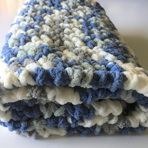 May include: A soft, crocheted baby blanket in shades of blue, gray, and white. The blanket is folded in thirds and has a fluffy texture.