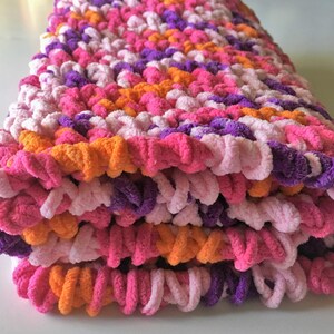 Puede incluir: Una manta de crochet rosa, morada y naranja doblada, hecha con un hilo esponjoso.