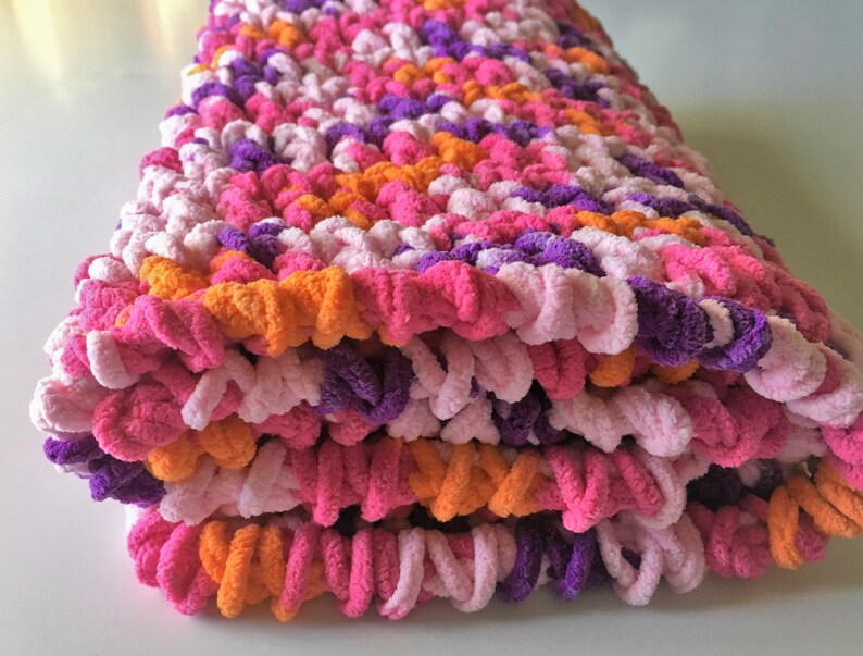 Puede incluir: Una manta de crochet rosa, morada y naranja doblada, hecha con un hilo esponjoso.