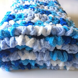May include: A blue, white, and light blue crocheted blanket folded in thirds. The blanket is made with a fluffy yarn and has a textured, bumpy surface.