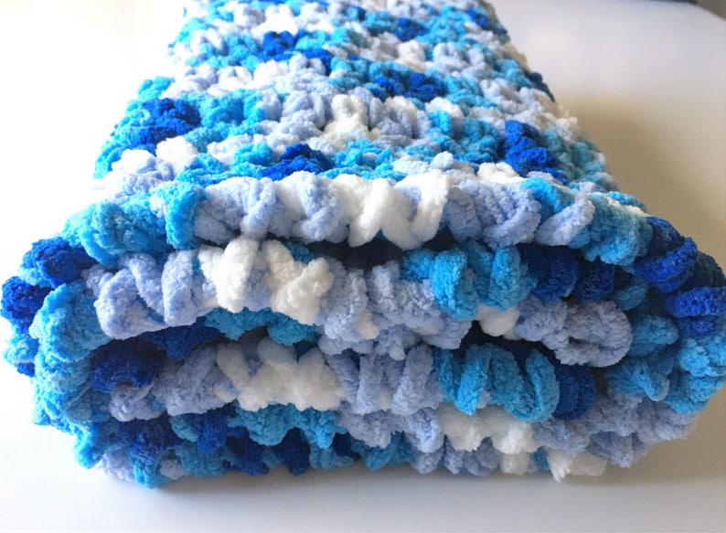 May include: A blue, white, and light blue crocheted blanket folded in thirds. The blanket is made with a fluffy yarn and has a textured, bumpy surface.