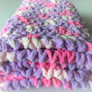May include: A soft, crocheted baby blanket in shades of pink, purple, and white. The blanket is made with a fluffy yarn and has a textured, chunky look.