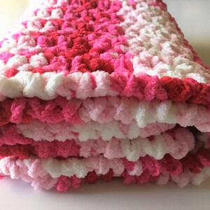 Puede incluir: Una manta de beb&eacute; de crochet rosa, blanca y roja doblada en tres. La manta est&aacute; hecha con un hilo esponjoso y tiene una superficie texturizada y abultada.