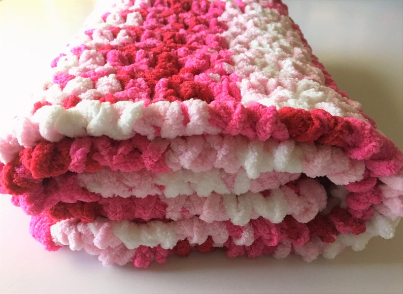 Puede incluir: Una manta de beb&eacute; de crochet rosa, blanca y roja doblada en tres. La manta est&aacute; hecha con un hilo esponjoso y tiene una superficie texturizada y abultada.