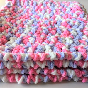 Puede incluir: Una suave manta de beb&eacute; tejida a crochet en tonos de rosa, blanco y lavanda. La manta est&aacute; hecha con un hilo esponjoso y tiene una puntada texturizada y gruesa.