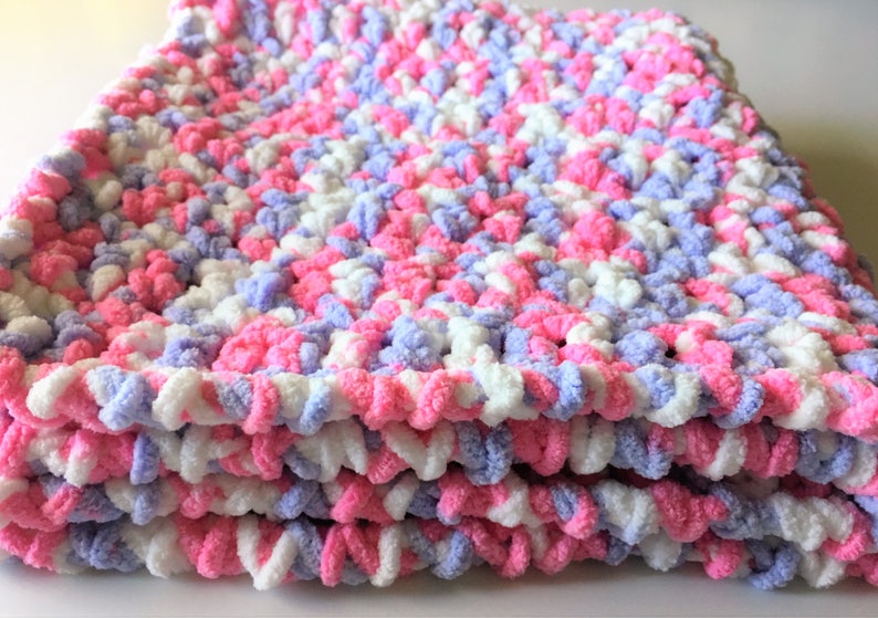 Puede incluir: Una suave manta de beb&eacute; tejida a crochet en tonos de rosa, blanco y lavanda. La manta est&aacute; hecha con un hilo esponjoso y tiene una puntada texturizada y gruesa.