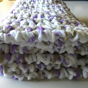 Puede incluir: Una suave manta de beb&eacute; tejida a crochet en blanco, morado y beige. La manta est&aacute; doblada en tres partes y tiene una textura esponjosa.