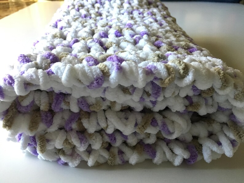 Puede incluir: Una suave manta de beb&eacute; tejida a crochet en blanco, morado y beige. La manta est&aacute; doblada en tres partes y tiene una textura esponjosa.