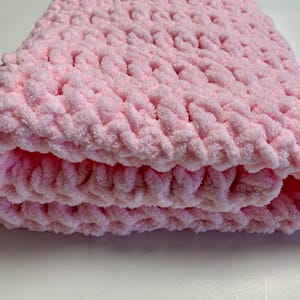 May include: A soft, pink crocheted baby blanket with a textured, fluffy yarn. The blanket is folded in thirds and is lying on a white surface.