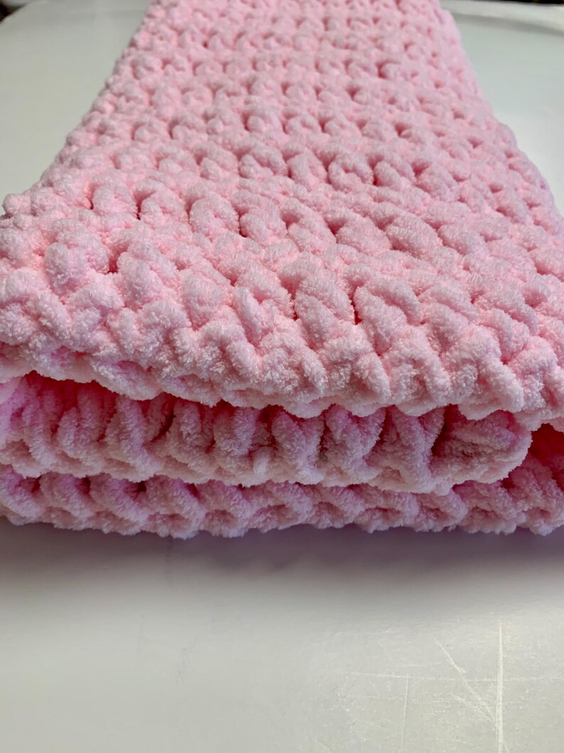 May include: A soft, pink crocheted baby blanket with a textured, fluffy yarn. The blanket is folded in thirds and is lying on a white surface.