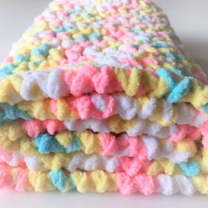 May include: A folded crocheted baby blanket in pastel colors of pink, yellow, and blue. The blanket is made with a soft, fluffy yarn.