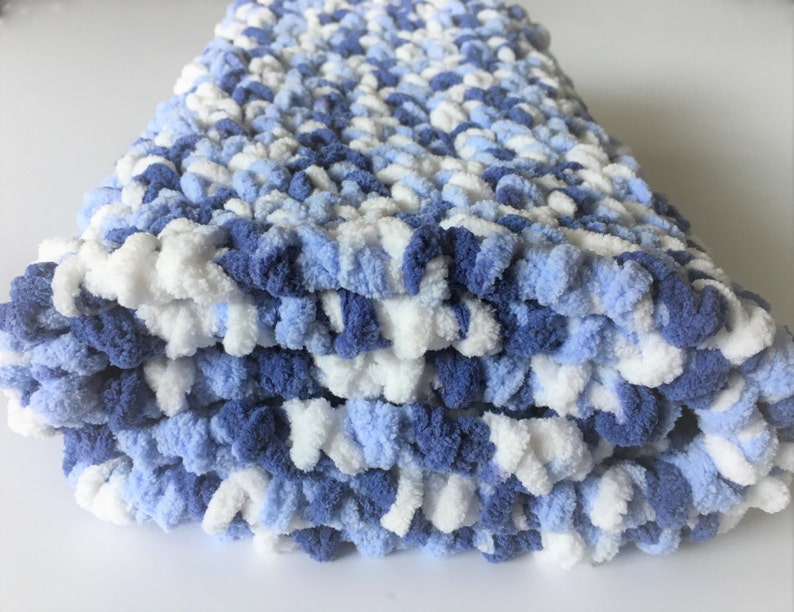 May include: A soft, blue and white crocheted baby blanket. The blanket is made with a chunky yarn and has a fluffy texture.