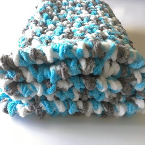 May include: A folded crocheted baby blanket in shades of blue, white, and gray. The blanket is made with a soft, fluffy yarn.